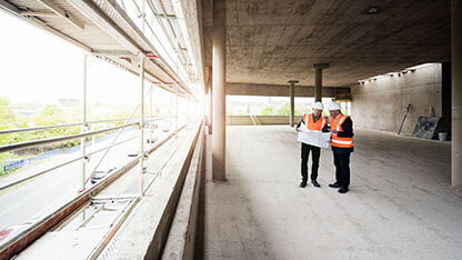 Two men with plan wearing safety vests talking in building under construction 