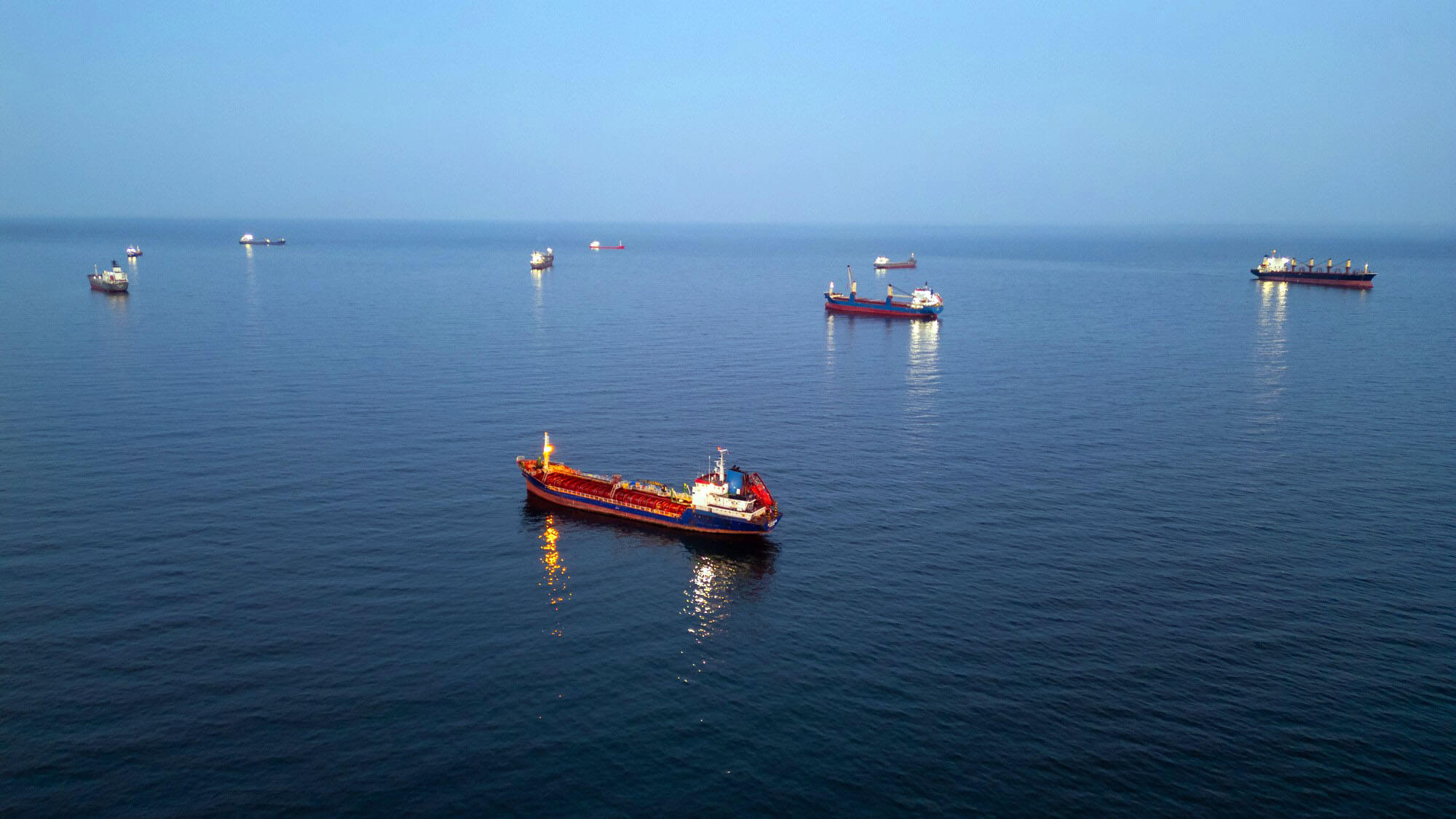 Cinematic drone shot of an oil tanker ship anchored at sea during twilight. The vessel is illuminated by onboard lights with a beautiful city skyline and sunset glow in the background.