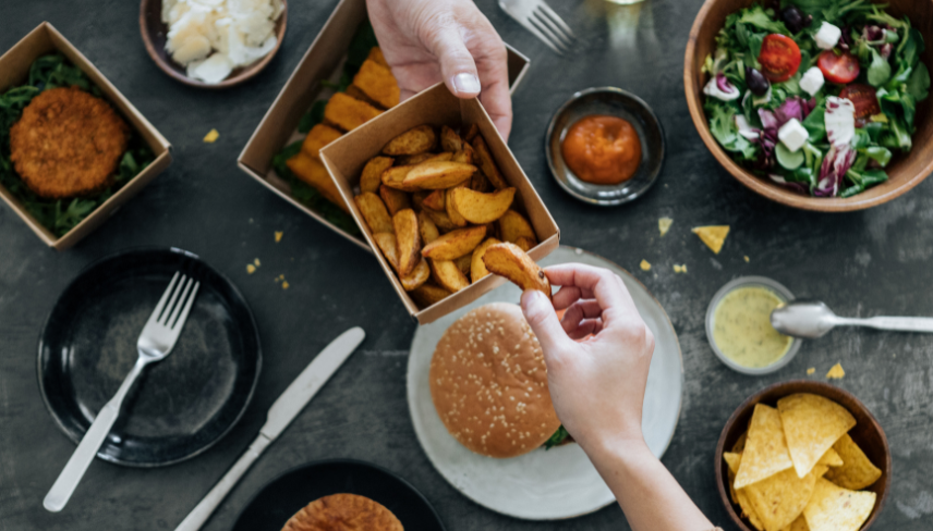 A person shares potato wedges from a box at a table set with a burger, salad, and other assorted foods.